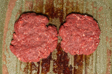 Two raw burger patties on a speckled green grill pan, with grease marks emphasizing the homemade preparation process before grilling.