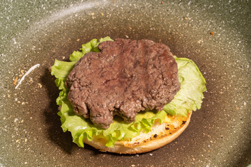 A cooked hamburger patty atop fresh lettuce on a toasted bun in a frying pan, capturing a rustic stage of homemade burger preparation.