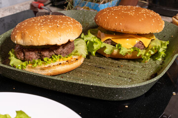 Two burgers with sesame buns, beef patties, lettuce, melted cheese, and sauce on a green grill pan, perfect for homemade meal scenes.