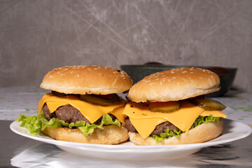 Two cheeseburgers with sesame buns, beef patties, melted cheddar, lettuce, pickles, and tomato sit on a white plate, ready for serving.