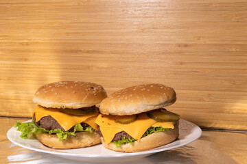 Two cheeseburgers with beef patties, cheddar cheese, pickles, lettuce, and tomatoes on sesame buns, served on a white plate with a rustic background.