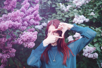 Teenage girl with red hair wearing glasses and blue blouse making a frame with her hands in front of blooming lilac bushes. Outdoor portrait photography
