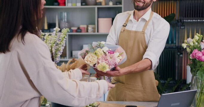 Woman buys flowers and pays with credit card using terminal. Male florist sells a beautiful bouquet to customer. Modern flower shop small business.