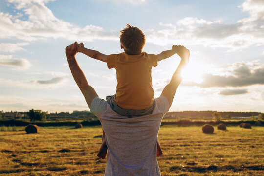 Father carrying son on shoulders in a sunny countryside field, holding hands and enjoying a joyful summer moment