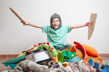 A boy sits on a pile of toys holding a sword and a shield with open arms.