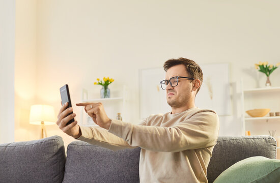 Serious man in glasses sitting on couch at home, holding smartphone at arm length, trying to discern. Young guy in eyewear squinting at screen, experiencing vision problems or blurred eyesight.