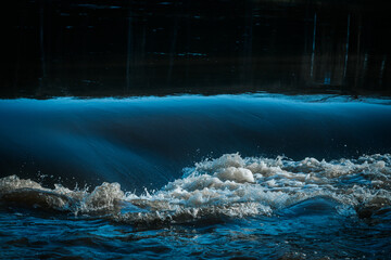 A powerful wave crashes in a river, with white foam and dark blue water creating a dramatic...