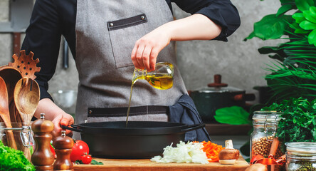 Chef pours olive oil in cast iron saucepan. Cozy kitchen with wooden table, kitchenware, vegetables, herbs and ingredients for cooking. Healthy food, eating, culinary, recipes, food blogging