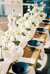 Black crockery stands on a wooden table next to white flowers and candles on a narrow tablecloth