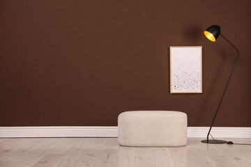 Stylish ottoman and lamp near brown wall in room. Space for text