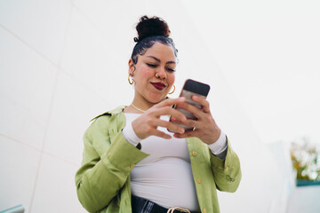 Fototapeta premium Stylish Latina with gold earrings focuses on her smartphone, tapping the screen with curiosity. Female is likely reading messages or engaging with content. Digital lifestyle and connectivity concept