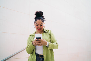 Young Latina in a green outfit looks at smartphone app, smiling as she types. Female seems engaged,...