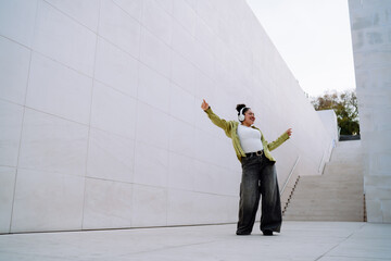 Smiling young woman listens to music while dancing in a modern architectural setting. Her energetic movements and stylish fashion reflect positivity, independence, and joy in everyday moments