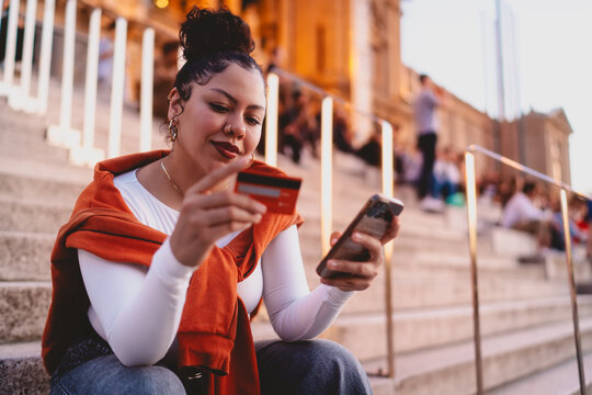 stylish young woman sits on outdoor steps, focused on making an online payment. Female holds credit card in one hand and smartphone in the other, enjoying digital shopping in a vibrant urban setting