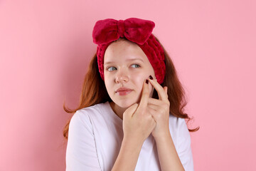 Teenage girl popping pimple on her face against pink background. Acne problem