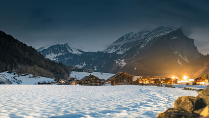 Winter night landscape in the mountains, snow-covered foothills with a small village in the Alps, houses illuminated at night, view from a hiking trail.