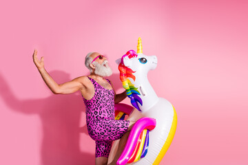 Elderly man enjoying summer fun in a pink leopard print bodysuit with a giant unicorn float on a...