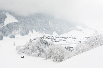 View of a small town in the Alps in a snowy mountain valley. Winter landscape with a view of a mountain hiking trail on a cloudy and foggy day.