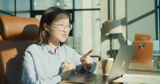 A woman in glasses having a video call in a modern office with a laptop and coffee, gesturing expressively and smiling under natural sunlight