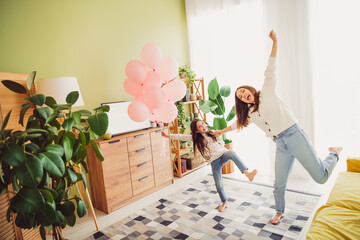 Joyful mother and daughter dancing in living room filled with sunlight and balloons on a cheerful...