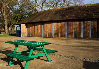 Green benches in the park with attached boards for playing chess, with a wooden building and trees in the background.
