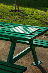 Green benches in the park with attached boards for playing chess, with trees and grass in the background.