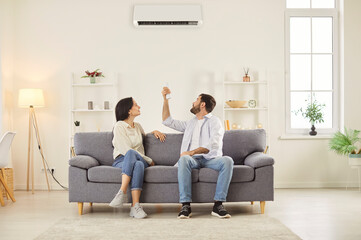 Married couple adjusts the air conditioner at home to control the climate. They ensure comfort by...