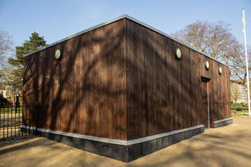A wooden building located in the park, with trees behind it. It’s a sunny day.
