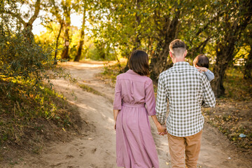 Happy family walking on the road in the park. Father, mother holding baby son on hands and going together. Rear view. Family Ties concept.