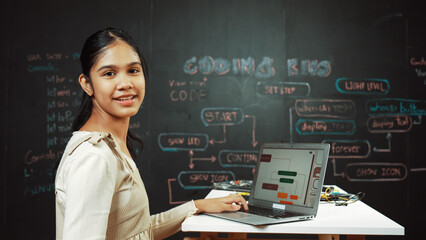 Caucasian girl working on laptop while smiling to camera at blackboard with engineering code and...
