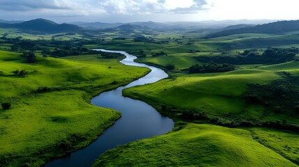 Meandering river flows through vibrant green rolling hills and valleys under dramatic cloudy sky, creating natural curves in pastoral landscape. Aerial perspective.