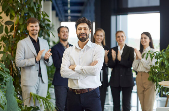 Portrait of a young happy smiling confident bearded business man in formal clothes looking cheerful at camera with a team of people company employees applauding in background in office.