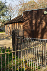 A fence in the foreground. A wooden building located in the park, with trees behind it. It’s a sunny day.