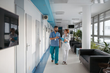 Two doctors walking across hospital hallway, holding medical records in hand.