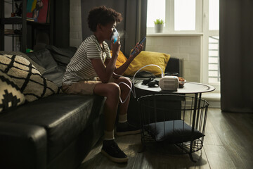 Child with asthma sitting on couch, inhaling fumes from inhalation mask and looking at smartphone...