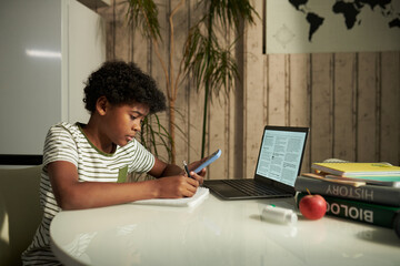 Side view of diligent schoolboy sitting by table in home environment and taking notes in copybook...
