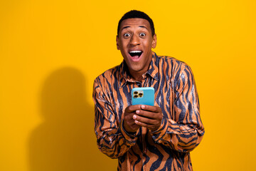 Excited young man using smartphone against yellow background wearing printed shirt and expressing...