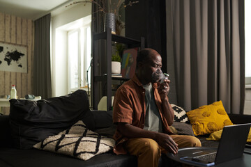 Senior man inhaling fumes from oxygen mask of nebulizer while sitting in front of laptop during...