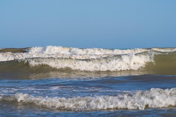 A light storm on the Spanish coast. Sea waves with crests of white foam roll onto a sandy beach on a clear day.
