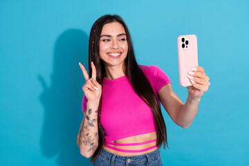 Young woman wearing a pink top takes a selfie, smiling and flashing a peace sign against a solid...