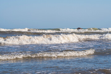 A light storm on the Spanish coast. Sea waves with crests of white foam roll onto a sandy beach on a clear day.