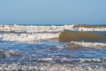 A light storm on the Spanish coast. Sea waves with crests of white foam roll onto a sandy beach on a clear day.