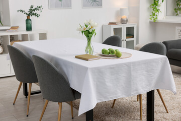 Table with white tablecloth, apples, flowers in vase and chairs in stylish room. Interior design