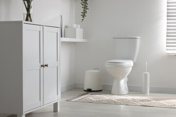 Toilet, brush and trash bin near white wall in bathroom