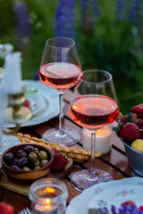 Romantic surprise, marriage proposal. Elegant decoration. Private wedding party for two, table set with floral decor, chandelier, fruits, wine in field or meadow. Summer, sunset, golden hour