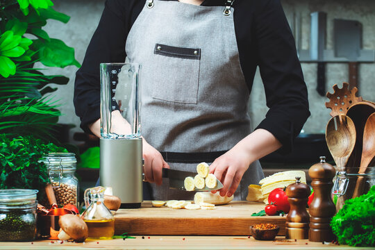 Chef  cuts bananas for prepare smoothie in blender. Cozy kitchen with wooden table, kitchenware, vegetables, herbs and ingredients for cooking. Healthy vegan eating, culinary, recipes, blogging