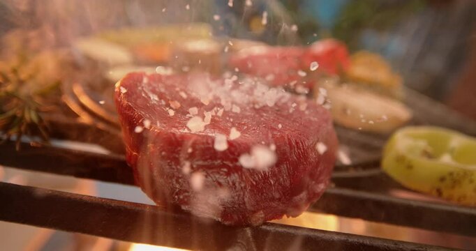 Panning down shot of an unrecognizable chef pouring sea salt on filet mignon while it is grilling on fire in slow-motion