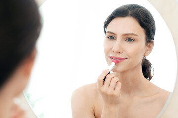 Pretty young woman looking at mirror, using lipstick, bedroom interior, empty space. Attractive...