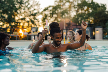 Friends enjoying drinks and dancing in swimming pool at sunset