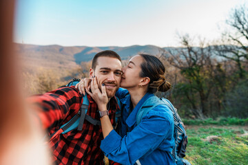 Young hikers couple taking a selfie while kissing on mountain top, enjoying the view of the valley...